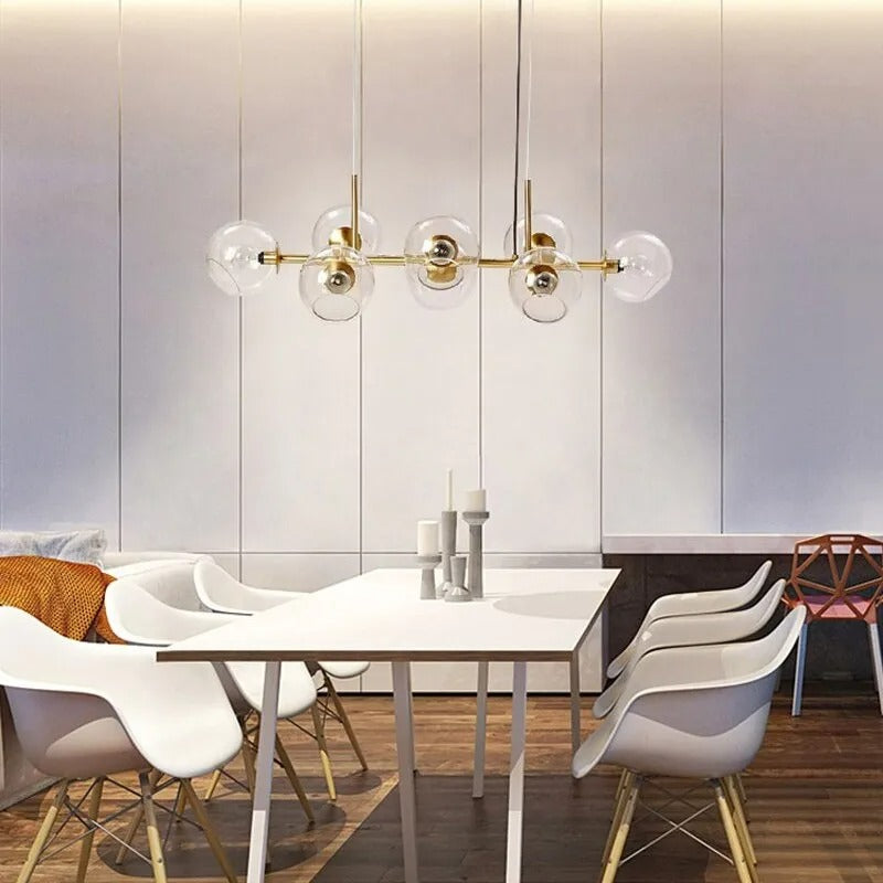 Gold Clérachic bubble glass linear pendant light above a white dining table set with wooden chairs.