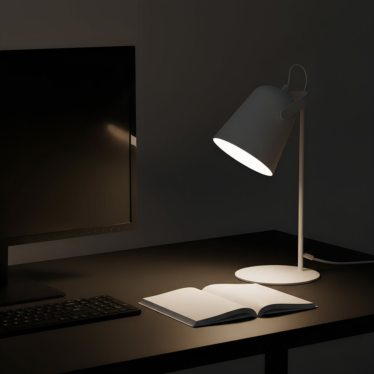 The white desk lamp on a working desk in the evening beside a dark monitor, its shade tilted down over an open notebook