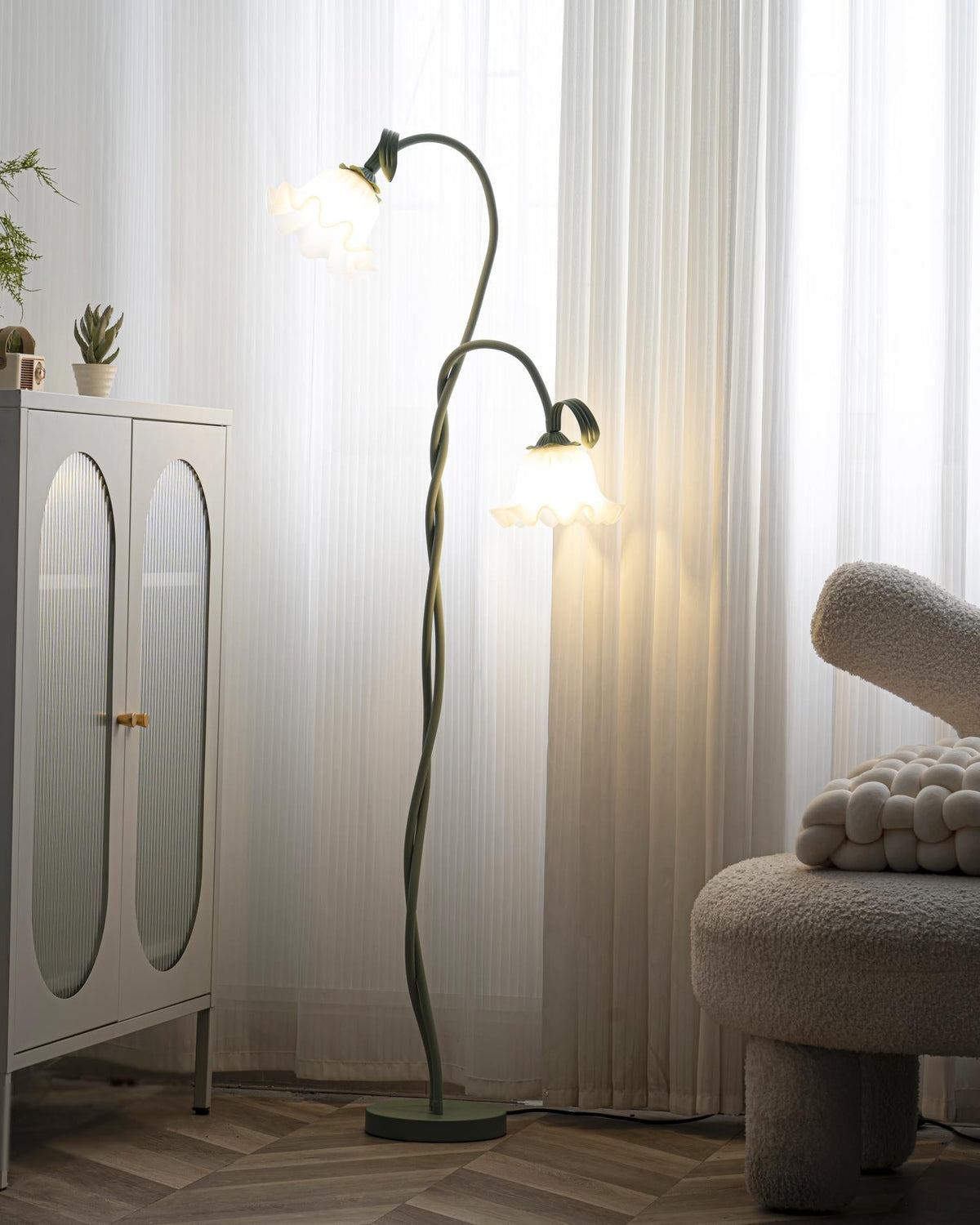 Clérachic green calla-flower floor lamp shown full length between a white cabinet and a bouclé chair, its lower bloom lit against sheer curtains.