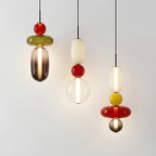 Three Clérachic pendants lit side by side on a pale wall, one stacking a red ball and olive disc over a smoked glass capsule.