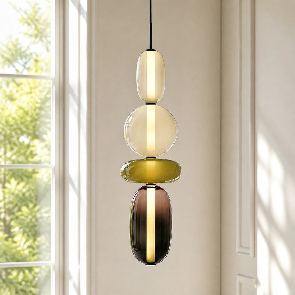 A tall Clérachic pendant glowing beside a sunlit window, its opal capsule and globe above an olive disc and smoked capsule.