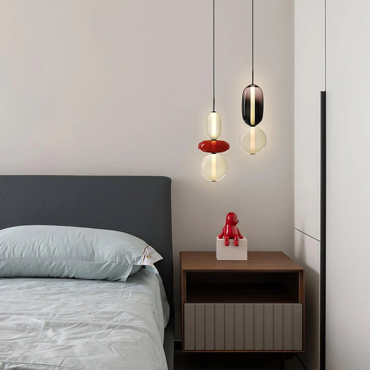 Two Clérachic pendants lit beside a bed with grey linen, one with a red disc and one with a smoked glass capsule.