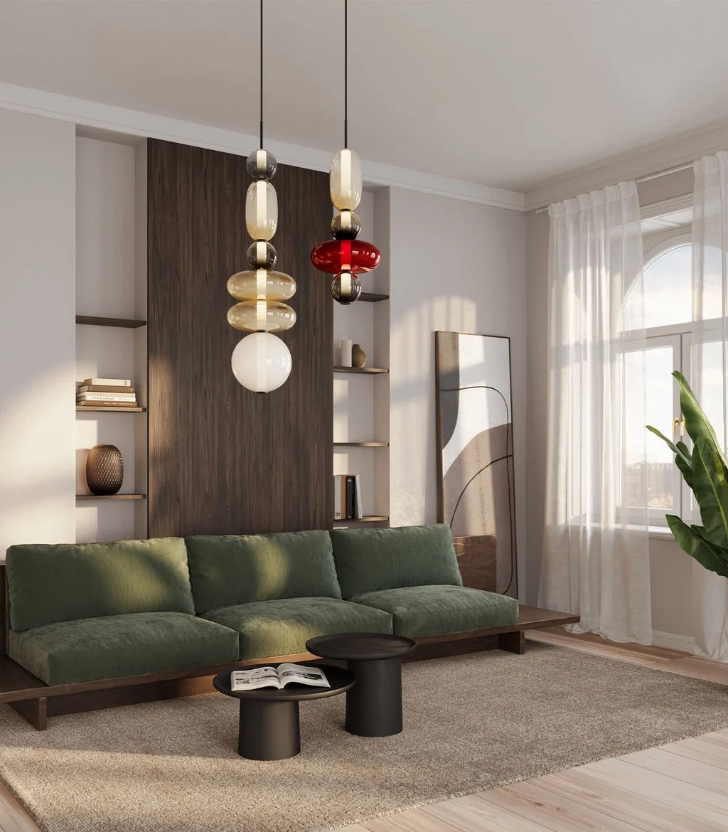 Two Clérachic stacked glass pendants, one in smoke, amber and opal glass and one with a red disc, hanging beside a green velvet sofa in a sunlit living room.