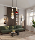 Two Clérachic stacked glass pendants, one in smoke, amber and opal glass and one with a red disc, hanging beside a green velvet sofa in a sunlit living room.