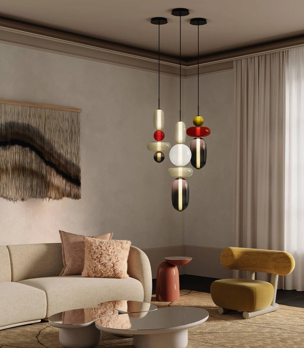 Three Clérachic stacked glass pendants in opal, red, olive and plum glass on black ceiling plates, lit above a cream curved sofa.