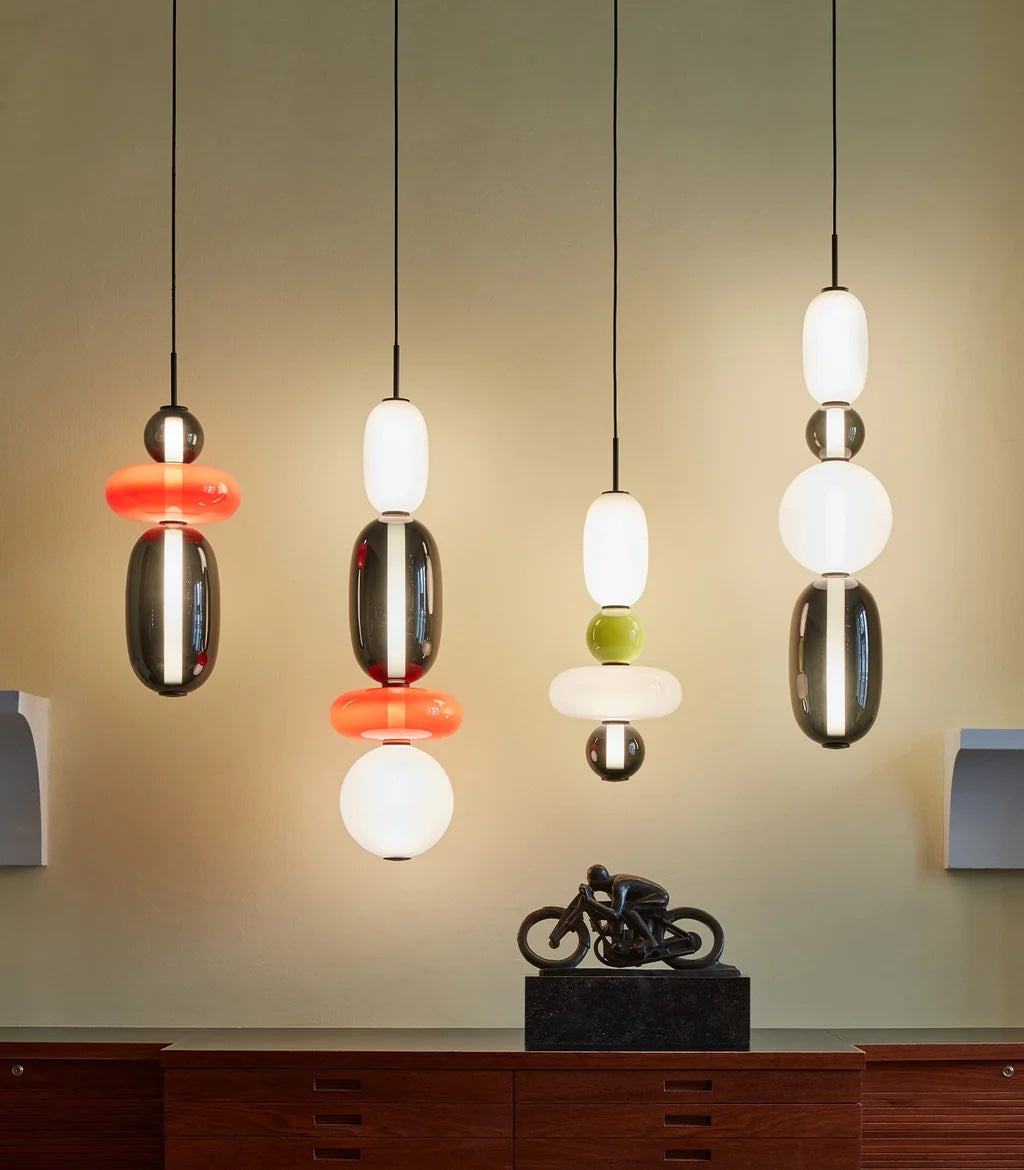 Four Clérachic stacked glass pendants in smoke, coral, opal and olive glass, lit against a pale green wall above a wooden sideboard.