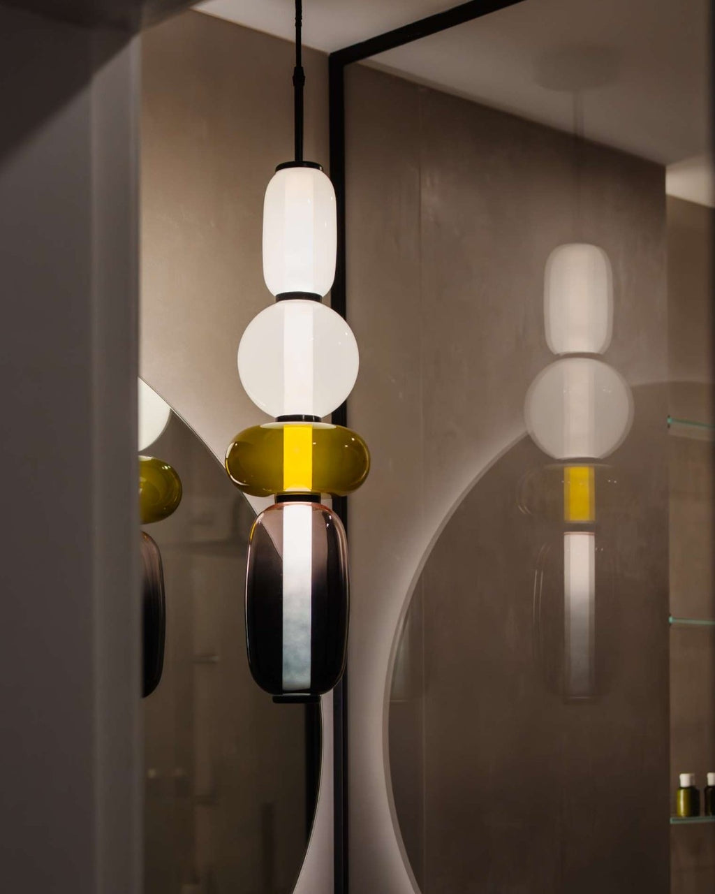 A Clérachic pendant of opal, olive and plum glass lit in a bathroom, its reflection showing in a round backlit mirror.