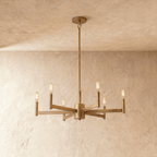 Clérachic six-arm chandelier in an aged brass finish with warm filament bulbs on bar arms, on a rod, against a limewash wall.