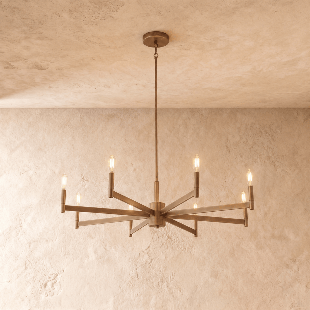 Clérachic eight-arm chandelier in aged brass with filament bulbs on straight bar arms, hung from a rod on a limewash backdrop.