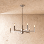 Clérachic six-arm chandelier in champagne gold with filament bulbs on square-section arms, hung from a rod on a limewash wall.