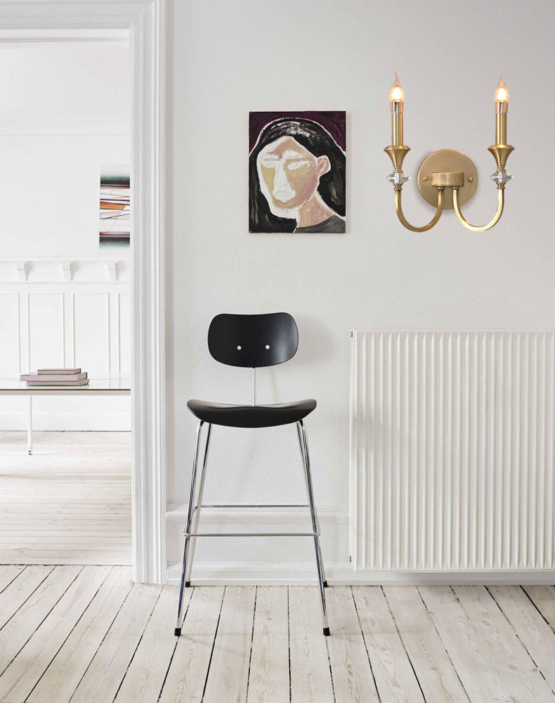 Small gold two-arm Clérachic wall light on a pale wall beside a framed portrait, with a black bar stool and radiator below.