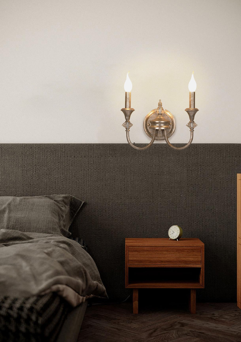 Gold two-arm Clérachic wall light above a walnut bedside table, glowing against a dark panelled wall in a bedroom.