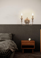 Gold two-arm Clérachic wall light above a walnut bedside table, glowing against a dark panelled wall in a bedroom.