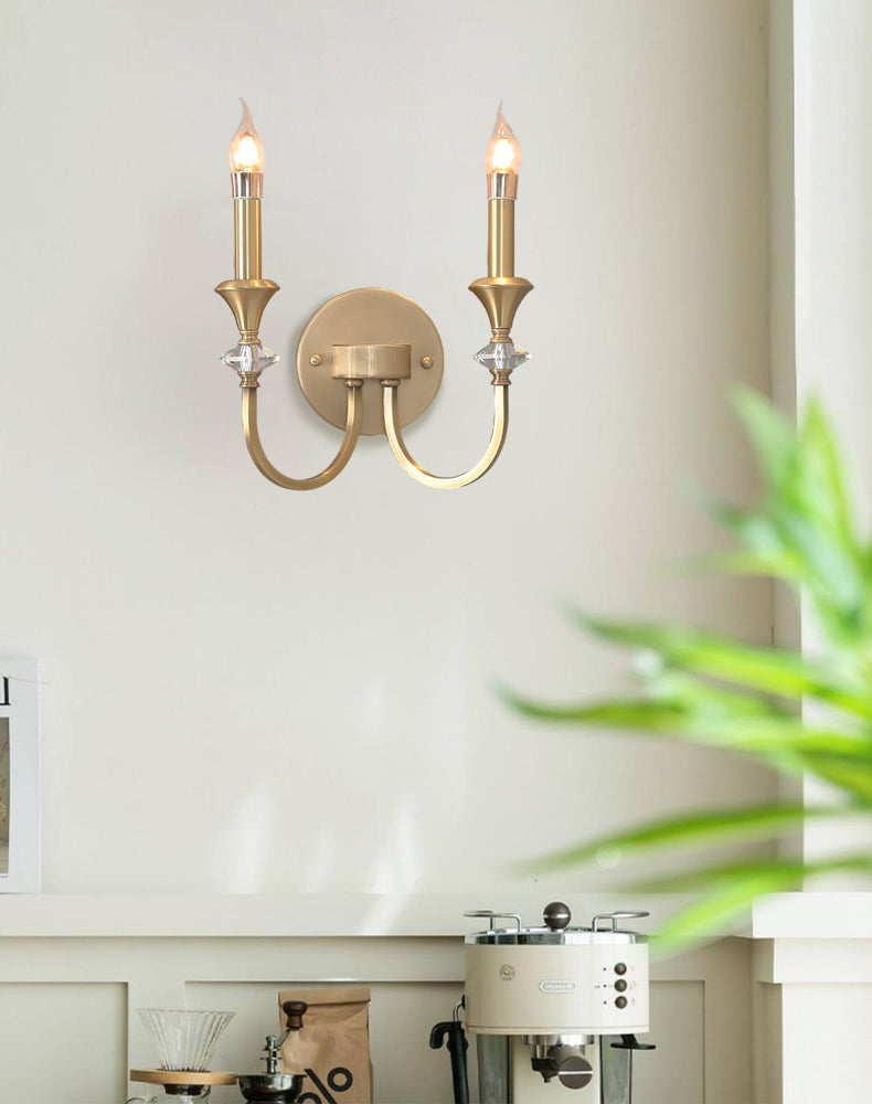 Gold two-arm Clérachic wall light on a pale wall, with a green plant at the right and a coffee machine on the counter below.
