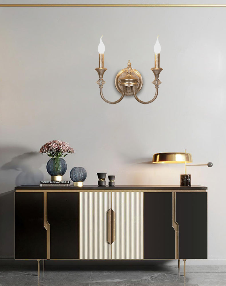 Gold two-arm Clérachic wall light centred above a black and cream sideboard, with a brass desk lamp and flowers on top.