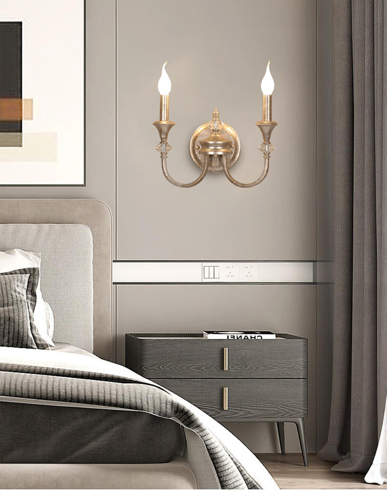 Gold two-arm Clérachic wall light on a grey wall above a dark bedside chest, with curtains at the right and a bed in the corner.