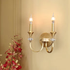 Lit gold two-arm Clérachic wall light on a warm cream wall, with red-pink blossoms in the lower corner.