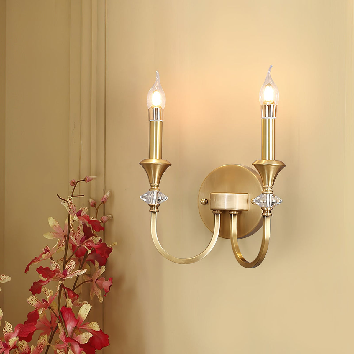 Lit gold two-arm Clérachic wall light on a warm cream wall, with red-pink blossoms in the lower corner.