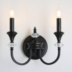 Front view of a black two-arm Clérachic wall light with two candle bulbs lit, mounted on a pale wall with crystal details.