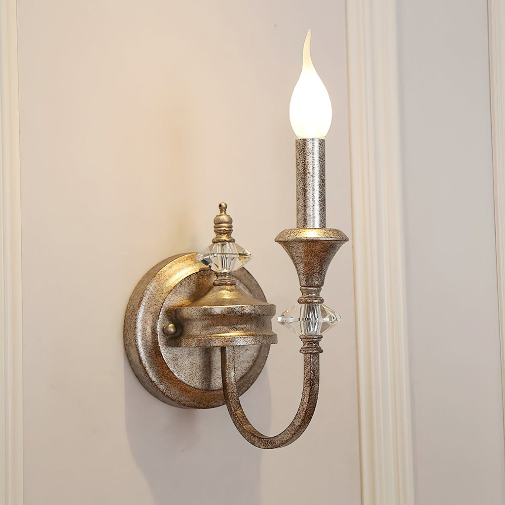 Single-arm Clérachic wall light in a worn rusty silver finish, its candle bulb lit against a cream panelled wall.