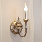 Single-arm Clérachic wall light in a worn rusty silver finish, its candle bulb lit against a cream panelled wall.