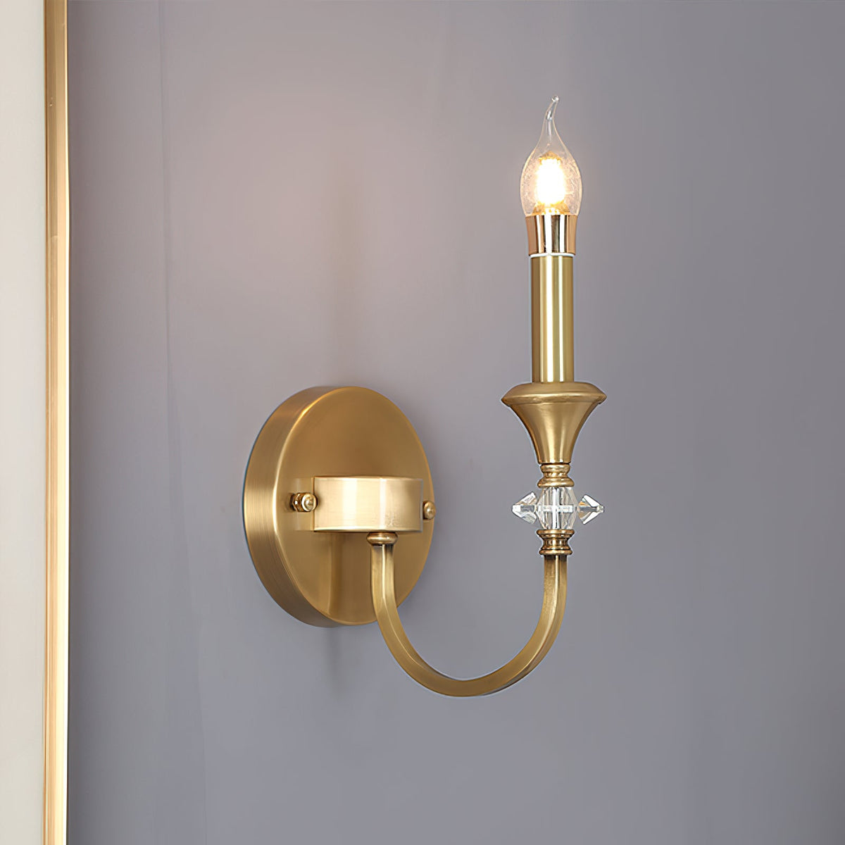 Gold single-arm Clérachic wall light on a dusty violet-grey wall, with a brass candle holder and a crystal detail on the arm.