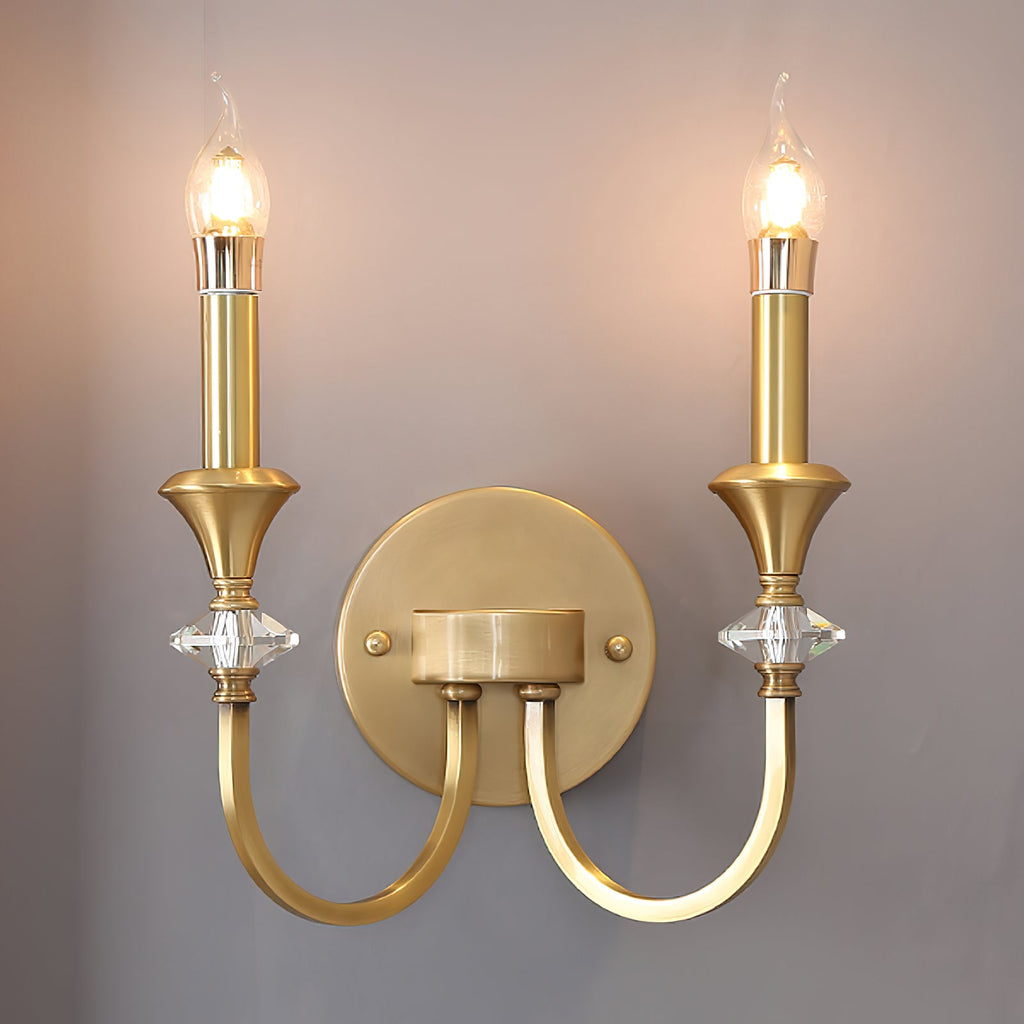 Front view of a gold two-arm Clérachic wall light with both candle bulbs lit against a muted mauve wall.