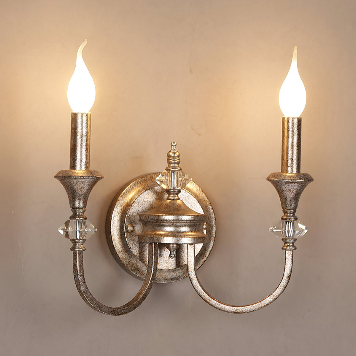 Rusty silver two-arm Clérachic wall light with both candle bulbs lit, set against a warm beige wall.