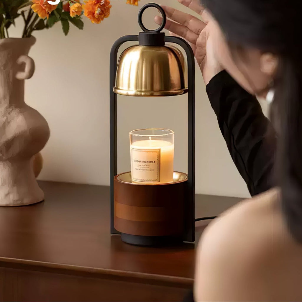 A woman reaches towards the gold dome of this Clérachic candle warmer lamp, a cream candle beneath it, on a dark wood console.