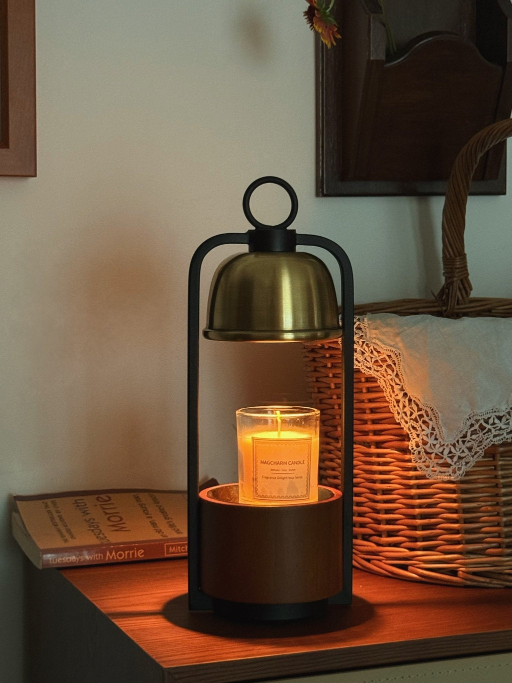 Clérachic candle warmer lamp lit on a bedside chest, the amber candle glowing under a gold dome, with a wicker basket beside it.