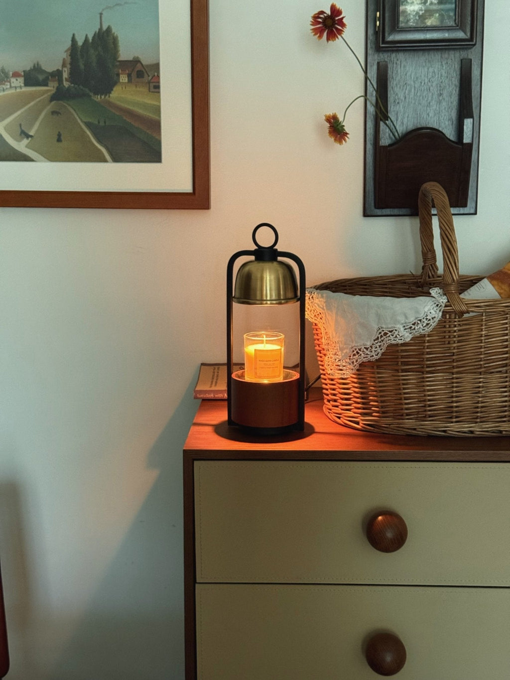 Small Clérachic candle warmer lamp lit on a cream chest with round wooden knobs, beside a wicker basket and a framed landscape.