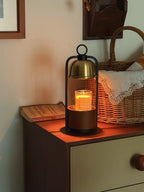 A Clérachic candle warmer lamp with its black arch and gold dome, candle lit, on a bedside chest in front of a woven basket.