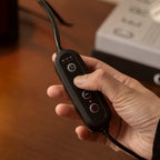 A hand holds the small black corded switch of this Clérachic candle warmer lamp, with a timer symbol and buttons on its face.