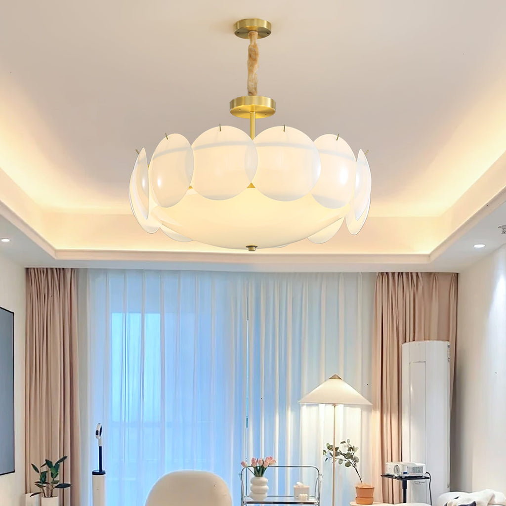 A lit Clérachic layered shell chandelier in a living room with a stepped cove ceiling, pink-beige curtains and a pleated floor lamp behind.