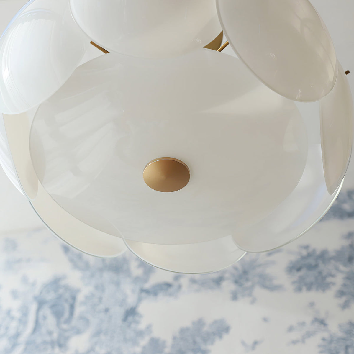 Looking up at the underside of this Clérachic chandelier, with a brass centre disc below the layered white shades, against blue toile wallpaper.