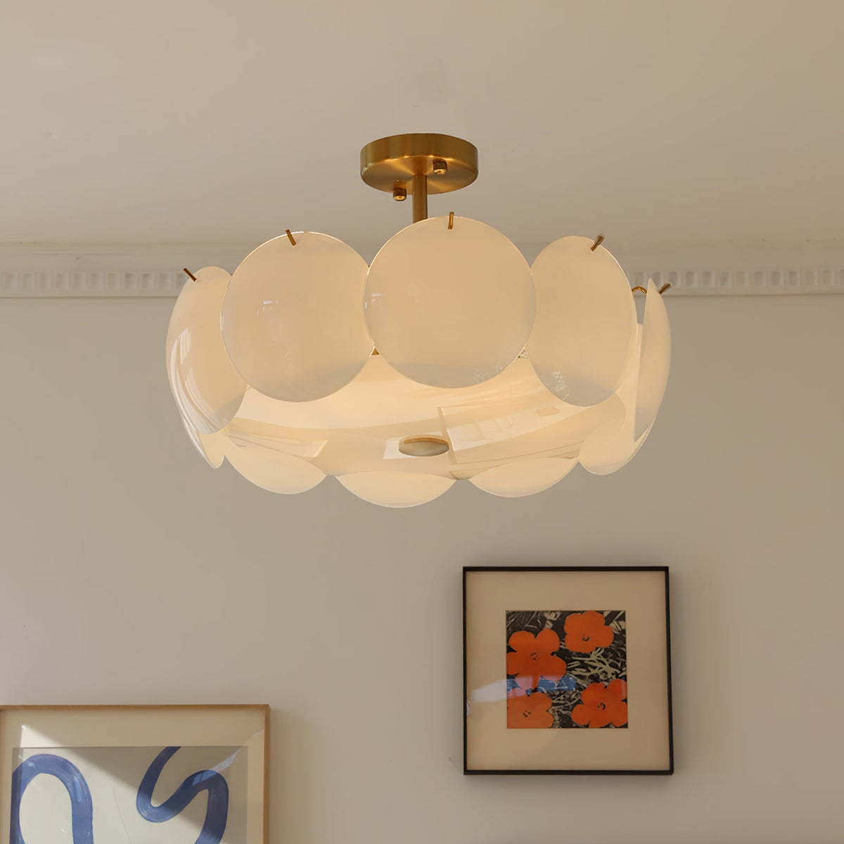 Lit Clérachic chandelier on a short brass stem, its ring of round white shades set in front of a cornice and two framed prints.
