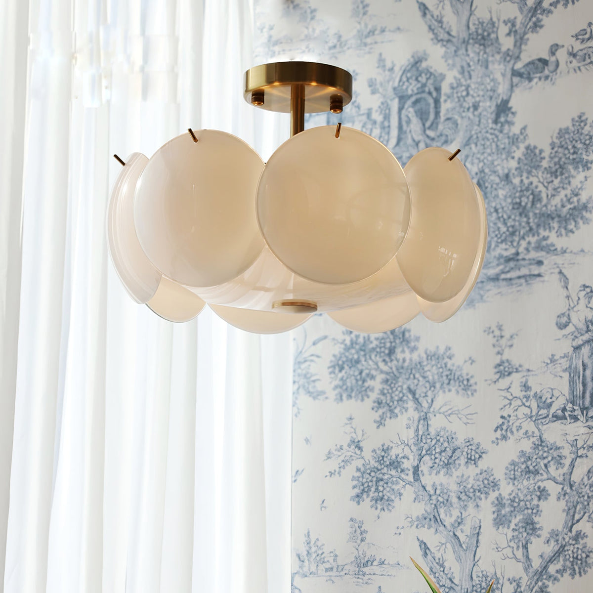 A Clérachic shell chandelier with a brass canopy, its shades warmed by light, in front of blue toile wallpaper and white curtains.
