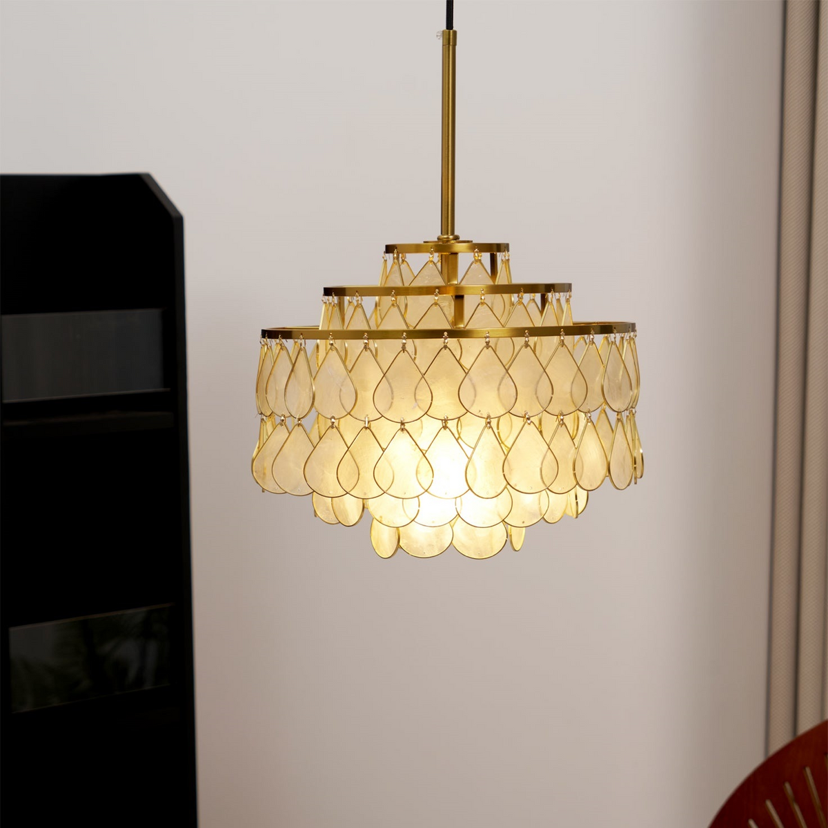 A small Clérachic chandelier hangs from a gold rod against a pale wall, its stacked rings of shell teardrops lit warm from within.