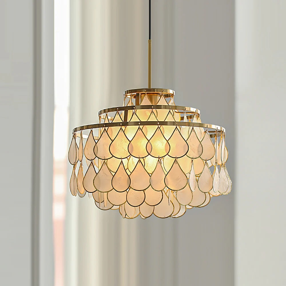Against pale sheer curtains, this Clérachic chandelier glows warm, three gold rings holding overlapping teardrop capiz shell discs.