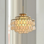 Against pale sheer curtains, this Clérachic chandelier glows warm, three gold rings holding overlapping teardrop capiz shell discs.
