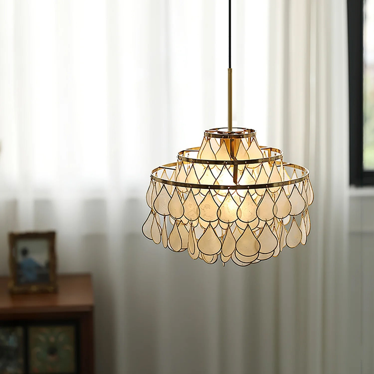 In front of long white curtains and a small framed photo, this Clérachic chandelier glows softly through its shell teardrops.