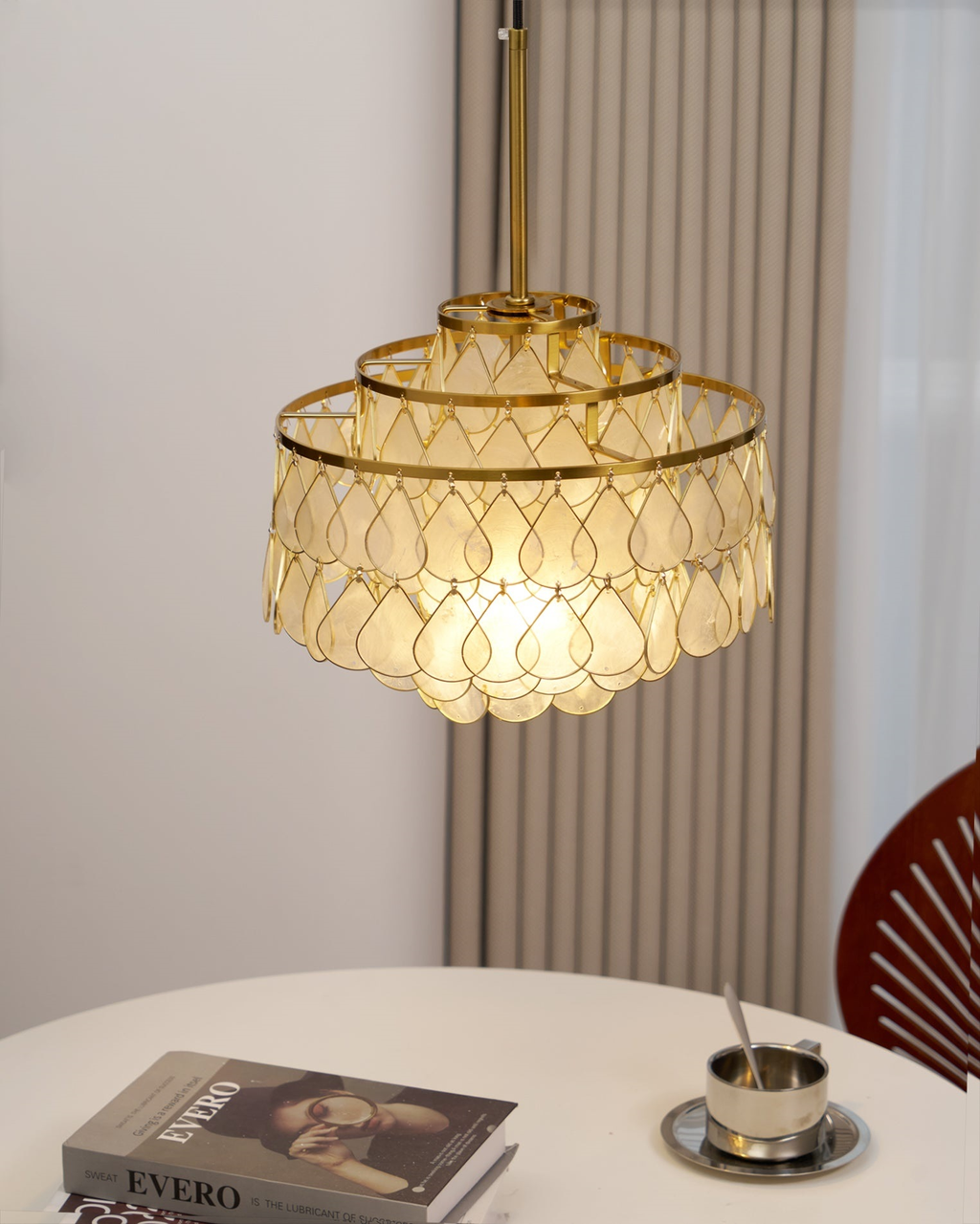 Above a round table with books and a coffee cup, this Clérachic chandelier hangs from a gold rod beside a ribbed curtain.