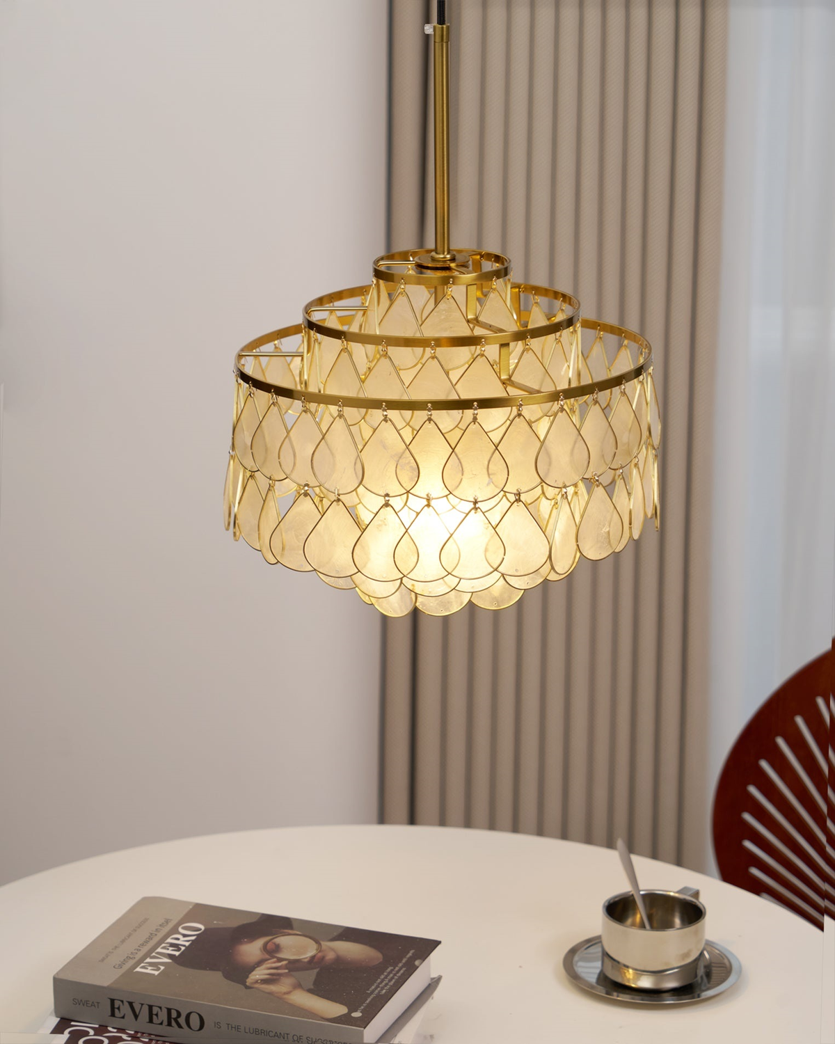 Above a round table with books and a coffee cup, this Clérachic chandelier hangs from a gold rod beside a ribbed curtain.
