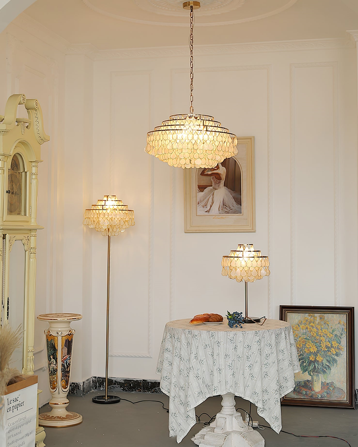 In a white panelled room with a grandfather clock and a lace-covered table, a Clérachic chandelier hangs above a matching floor and table lamp.