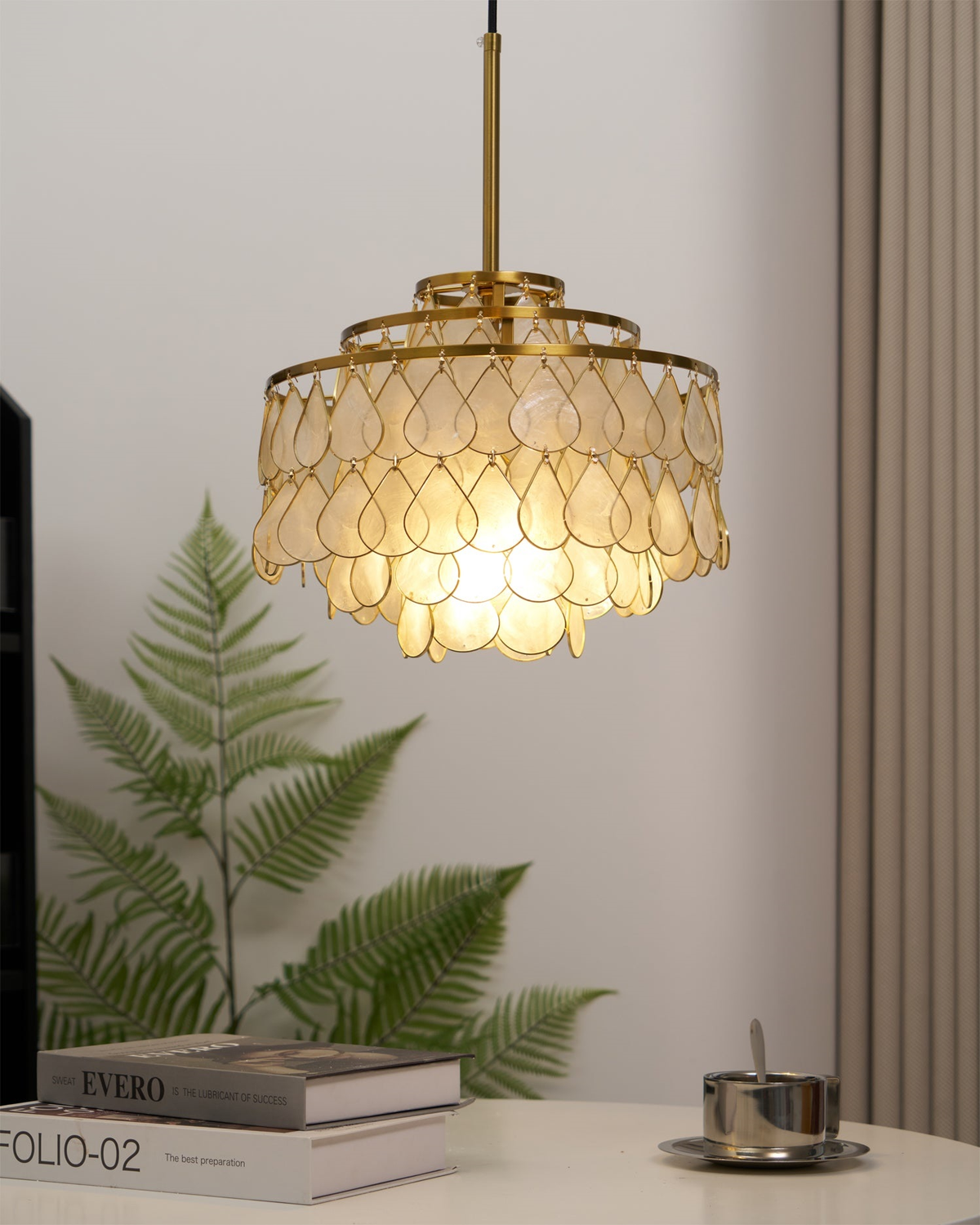 Beside a fern and a stack of books, this Clérachic chandelier hangs from a gold rod, its shell teardrops glowing a soft warm gold.