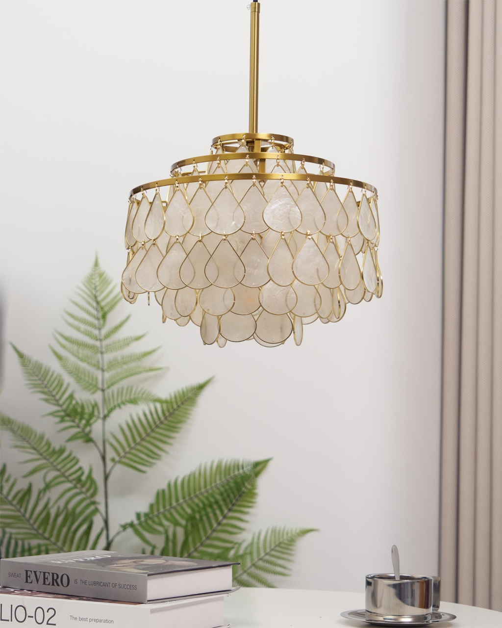 A Clérachic chandelier on a gold rod in front of a fern and a pale grey curtain, its shell teardrops in unlit daylight.