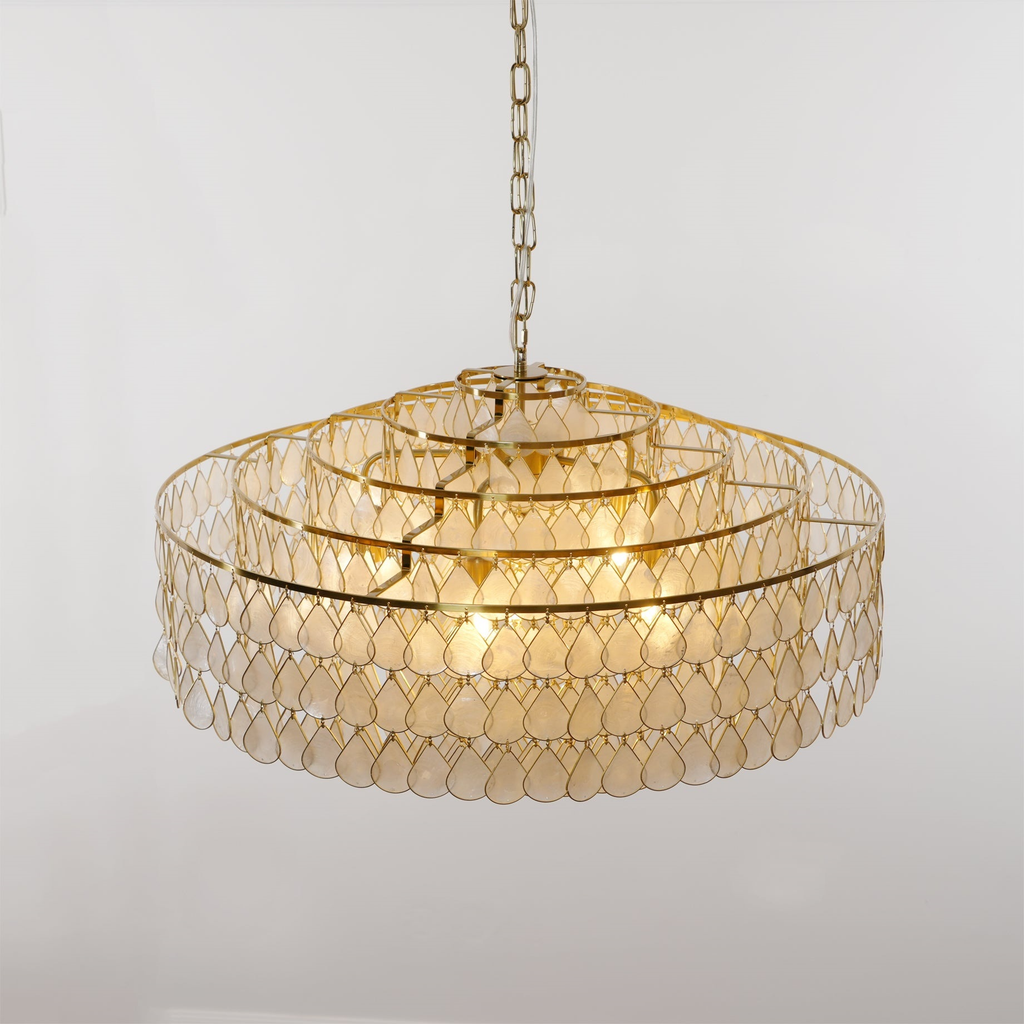 A wide Clérachic chandelier on a short chain, its four gold rings stacked above capiz shell teardrops, shown on a pale grey wall.
