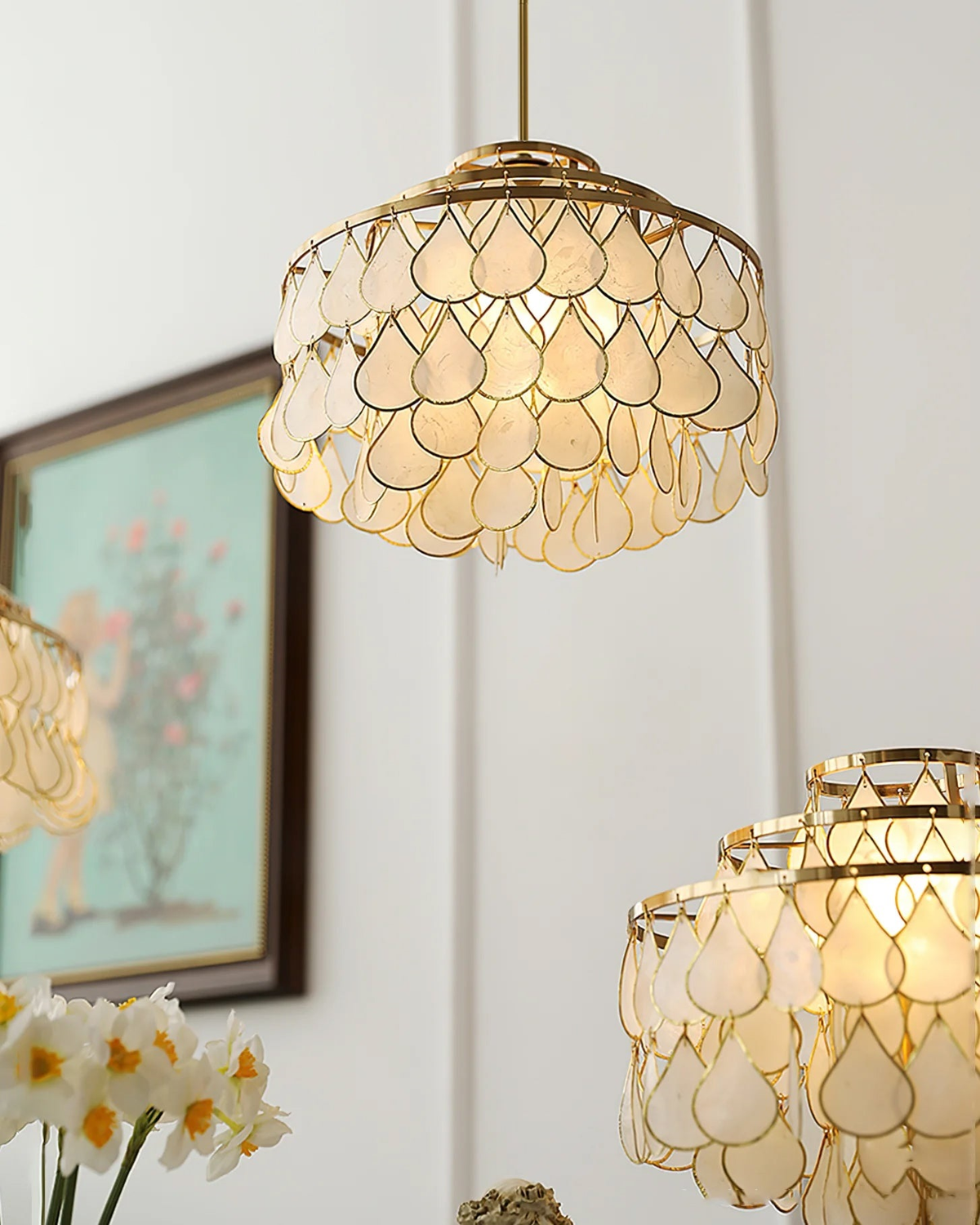 Seen from below, a Clérachic chandelier glows warm, its tiers of shell teardrops framed with an angel figure and a painting at the edge.