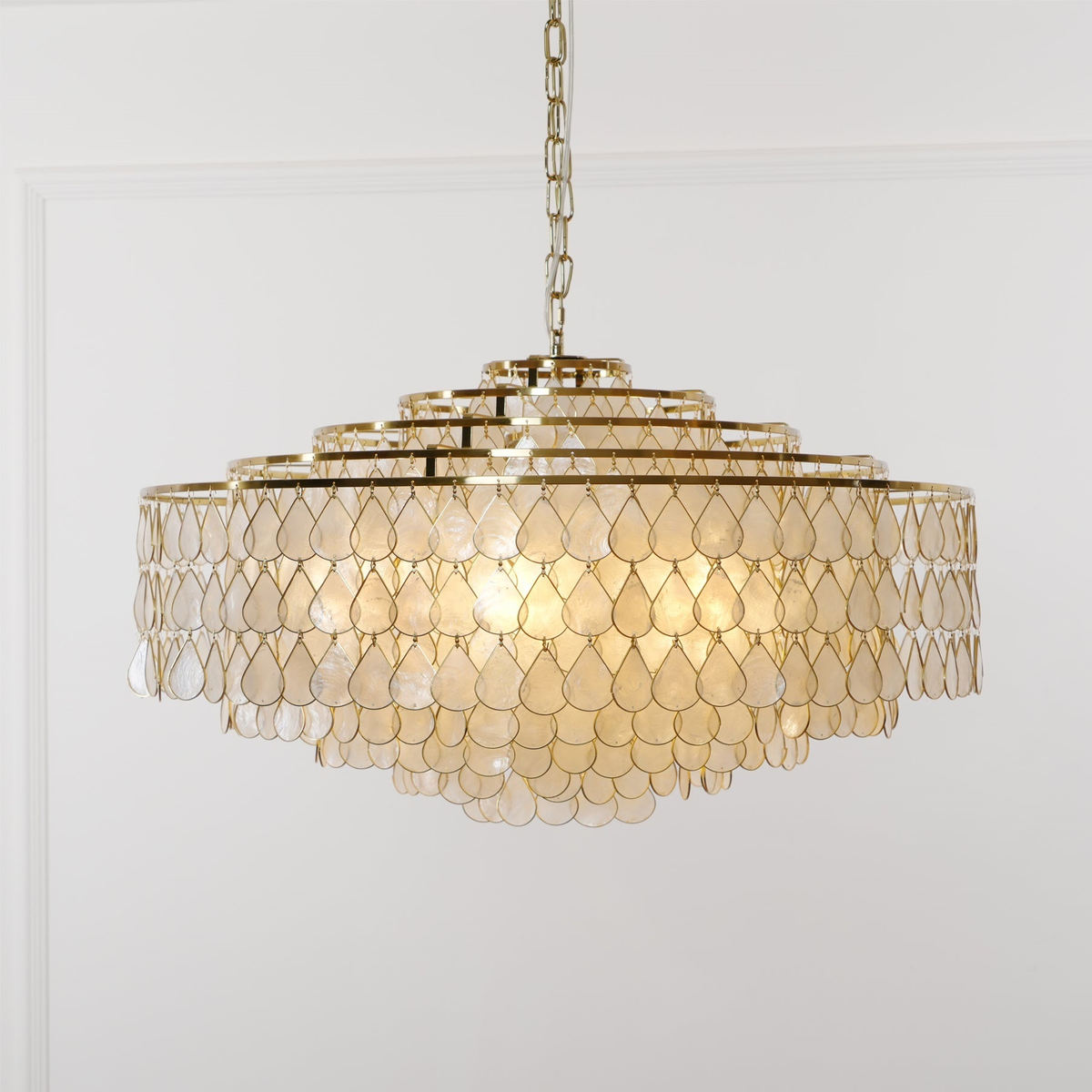 A big Clérachic chandelier on a chain, its tiered gold rings lit warm behind rows of teardrop capiz shell discs, in front of a panelled wall.