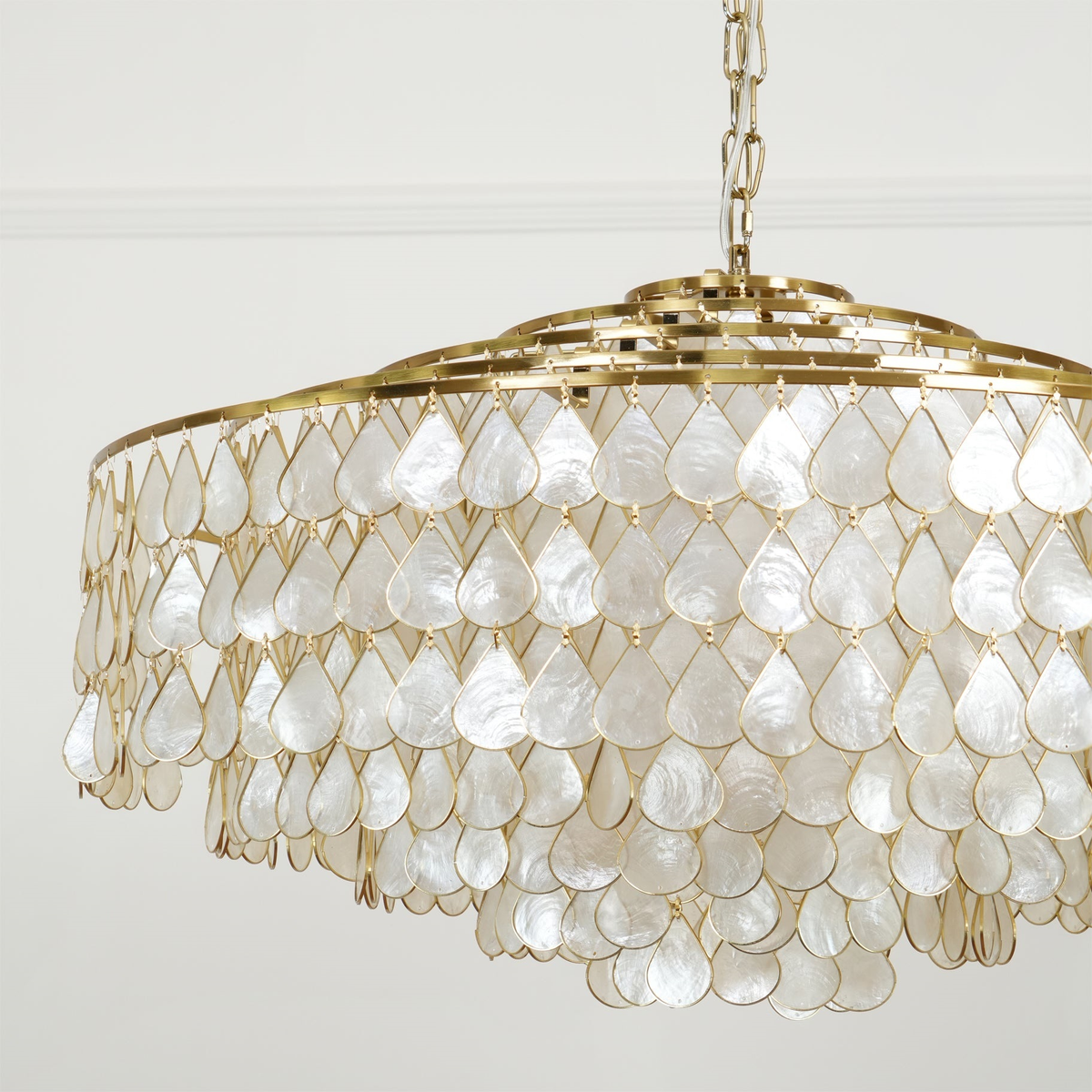 The pearly side of this Clérachic chandelier, with rows of white shell teardrops on gold rings, hung from a chain against a white wall.